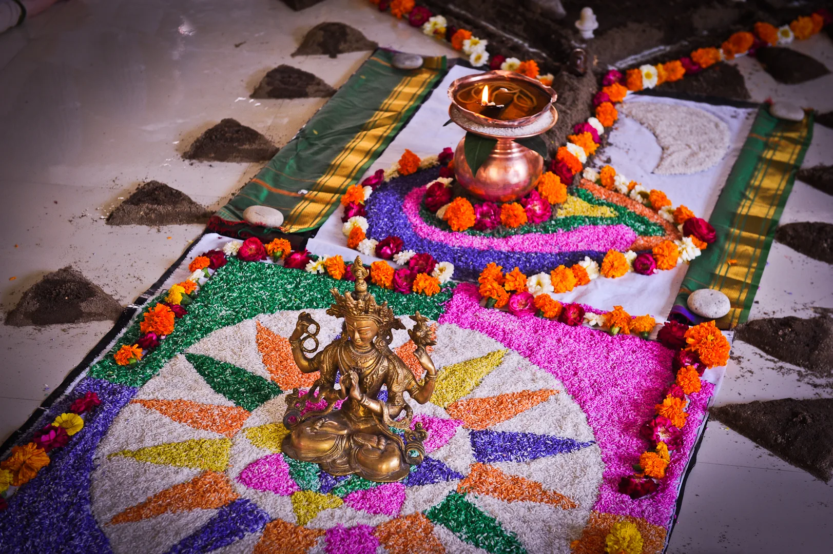 A puja altar: an oil lamp and a small brass deity on a yantra of coloured rice, framed with marigolds